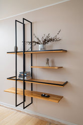 Solid wood floating shelf PÄIKE with two metal frames on a living room wall decorated with a plant and books